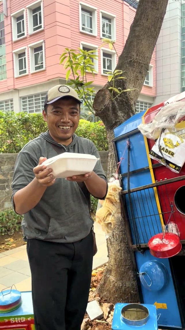 Pedagang keliling bertopi tersenyum sambil memegang nasi kotak di samping gerobaknya