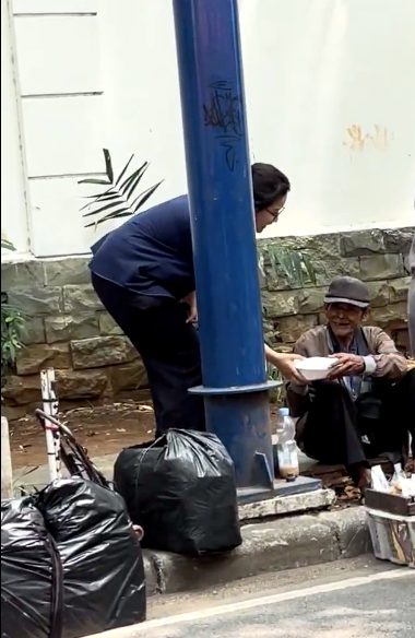 Relawan membungkuk menyerahkan nasi kotak kepada seorang bapak lanjut usia yang duduk di tepi jalan